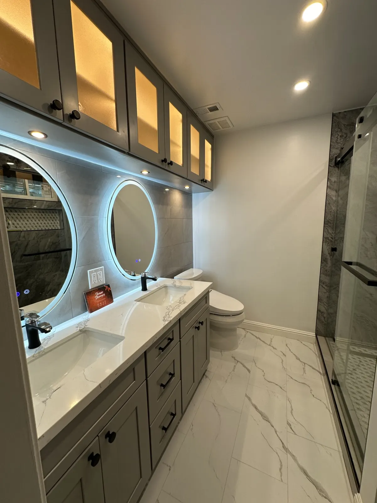 Bathroom with round backlit mirrors, a gray vanity, and a glass shower